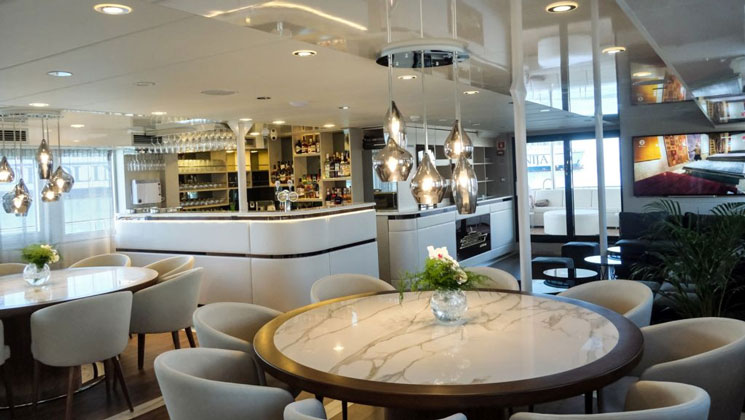 Dining room with white marble tables, beige padded chairs, white leather-wrapped bar & modern glass pendant lights on deluxe yacht Aurelia.