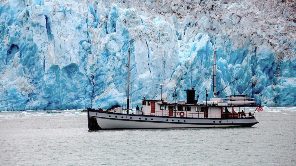 Classic wooden small ship navigates in front of a jagged teal icy Alaska glacier during a Glacier By National Park cruise.