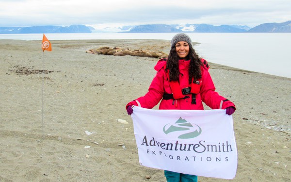 Single traveler in a red jacket holds an AdventureSmith flag on an Arctic beach