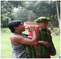 Person playing a conch shell and another man dressed in traditional Hawaii outfit with green headdress and lai