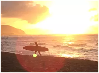 Surfer silhouette walking along Hawaiian beach with sun setting