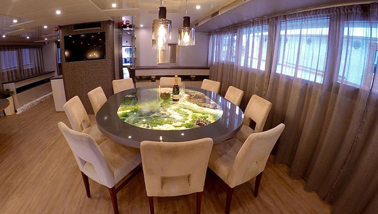 Dining room aboard yacht Rhapsody with art deco green marble round table, beige padded chairs, pendant lights, wood floor & windows.