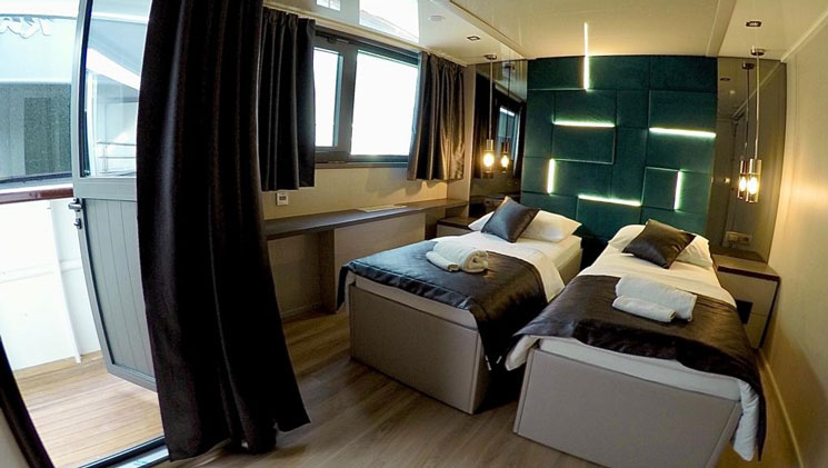 VIP Upper Deck cabin on yacht Rhapsody, with 2 twin beds, wood floor, dark curtains, lit up turquoise headboard & pendant lights.