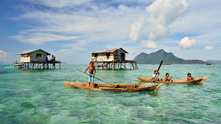 Kids in sea gypsy village Bajau Laut, Indonesia, on a wooden boat in front of houses on stilts over emerald water on a sunny day.