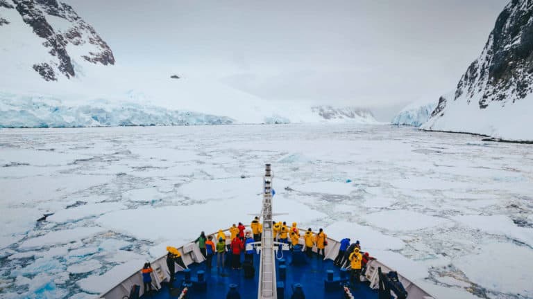 Antarctica travelers stand at bow of ship to view iceberg-laden waters & snowy mountains on a cloudy day.