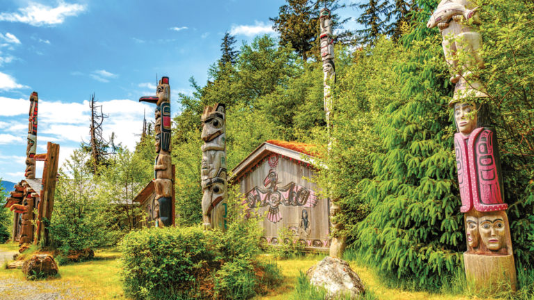 Native American Totems and Clan Houses in Ketchikan, Alaska, set against bright green forest and blue sky.