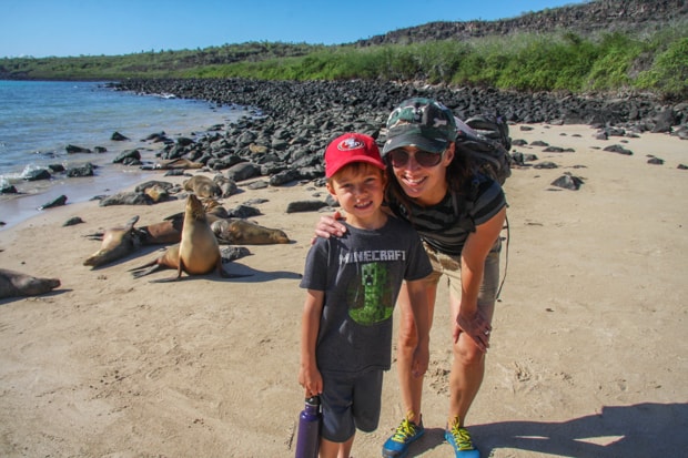 A mother in a blue hat and young son in a red hat standing in front of seals on the beach in Galapagos.