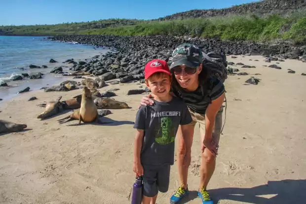 A mother in a blue hat and young son in a red hat standing in front of seals on the beach in Galapagos.