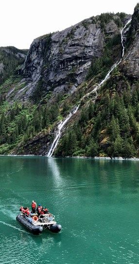 a daily excursion aboard an Alaska small ship cruise, a group enjoys a skiff ride through the teal waters of Alaska's Inside Passage, beyond them is a lush mountainside with a waterfall coming from the cliff. 
