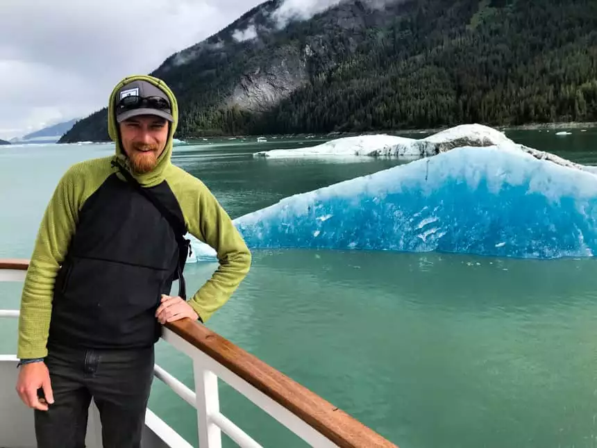 Adventure specialist on bow of alaska small ship in front of forested mountain range and bay water filled with floating teal icebergs
