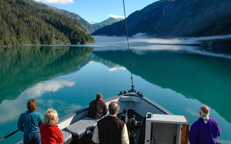 5 travelers gather on the bow of an Alaska small ship looking forwards at a glacier at the end of the green watered fjord, sandwitched between a mountain range, seen on Alaska's Eastern passages cruise