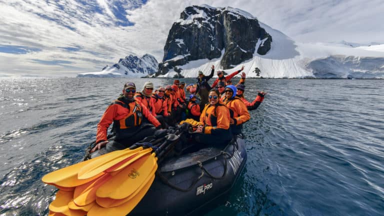 A group of Antarctica travelers in brightly colored jackets waves from a Zodiac with yellow kayak paddles on the bow, on a sunny day during the Antarctic Southern Latitudes expedition.