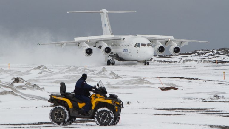 A man rides a yellow all terrain vehicle across Antarctic tundra, beyond him a white charter plane sits on a landing strip in Antarctica.
