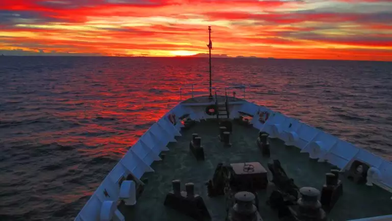 Small ship bow of white & green sails through calm seas under a blazing sunset of orange, yellow & pink in the Drake Passage.