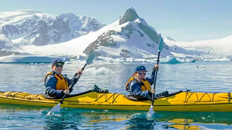 Kayakers paddle a yellow tandem kayak through icy blue waters with snowcapped peaks in the background on a sunny day in Antarctica.