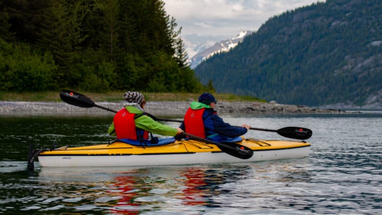 Tandem kayakers paddling on a Southeast Alaska small ship cruise, with green mountains and rocky shoreline in the background.