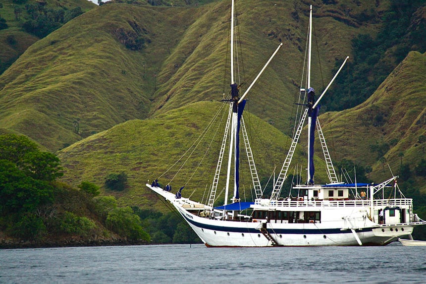 a white Indonesian wooden sailing ship against green lush mountain side floating in the ocean