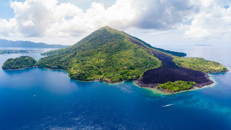 Aerial view of the Gunung Api volcano, covered in bright green vegetation & surrounded by deep blue sea, seen during the Aqua Blu Ambon & Spice Islands Cruise in Indonesia.