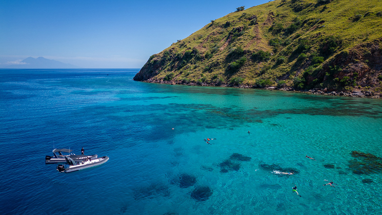 2 small tender boats sit at edge of turquoise sea as guests on Aqua Blu Raja Ampat Cruise snorkel beside bright green mountains.