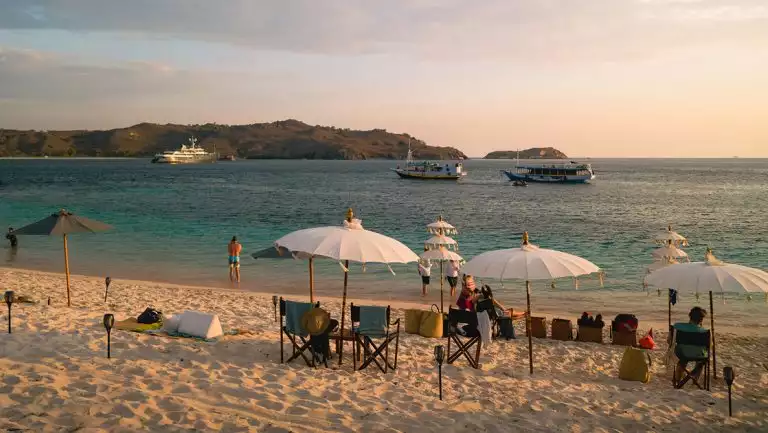 White-sand beach set up with ornate umbrellas & camping chairs by turquoise water with 3 small ships offshore at sunset.