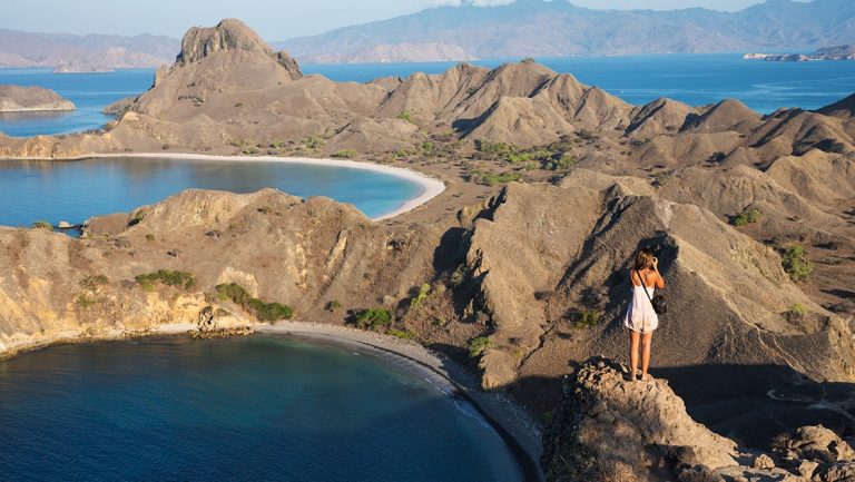 A female guest from a Komodo national park cruise stands on top of the jagged volcanic mountains to take a photo of the dramatic landscape.
