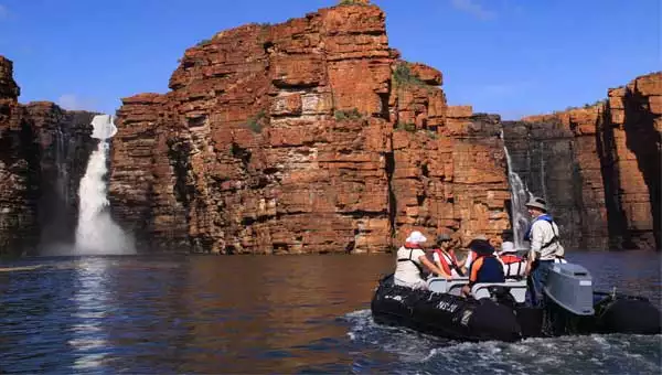 Travelers in a skiff explore red cliffs and a towering waterfall in the Kimberley of Australia