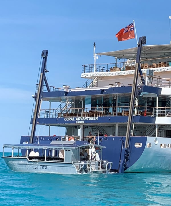 A photo from the water looking at the stern of the Coral Adventurer small Australian ship. The four levels of decks are shown with a multi level hydraulic lift. At the bottom of the lift is a smaller passenger vessel 