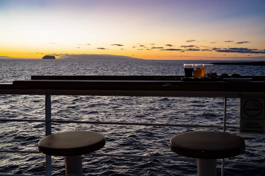 Stools and a high top table holding a selection of beers overlooking the back of a small ship with a view of the orange purple sunset