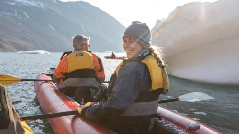 A woman paddler in a tandem sit-in-top, inflatable kayak, looks back and smiles on a sunny day, during the Greenland Adventure cruise by land, sea and air.