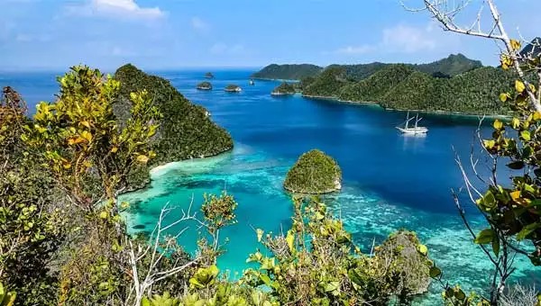 A remote bay in Raja Ampat Indonesia with a white, masted small cruise ship anchored at the shore.