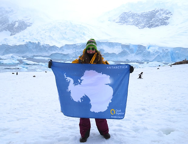 In front of an icy white landscape a female traveler wears a yellow expedition jacket and holds a blue and white flag of antarctica, she is celegrating reaching her 7th continent via Antarctica cruise