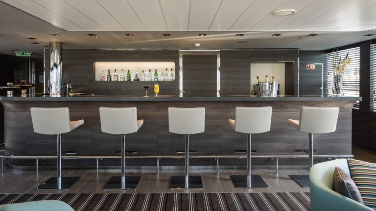 grey bar with 5 tall white leather bar seats at the counter in front are a couple teal accent chairs sitting ontop of a geometric grey scale rug aboard Le Soleal expedition ship