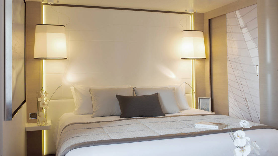 Deluxe Stateroom with king bed aboard Le Soleal luxury expedition ship, caramel colored walls with white bedding and headboard and featuring grey accent pillows