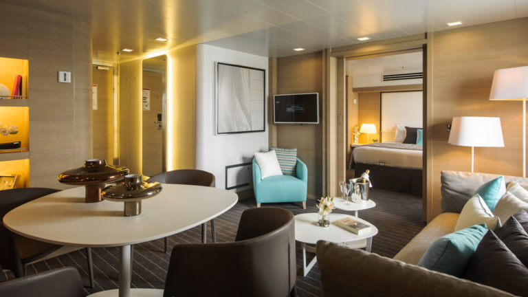 Owner's Suite aboard Le Soleal, two rooms interconnected, one with seating area and table the other with king size bed, the color decor is grey, tan and pops of teal