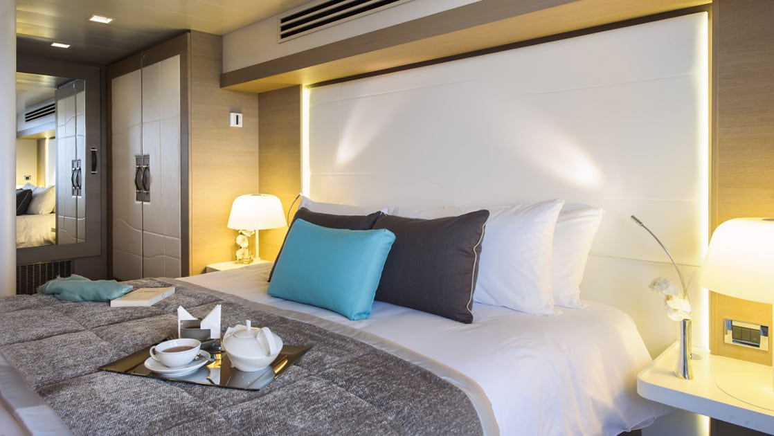 Prestige Stateroom aboard Le Soleal with king bed. A tall white headboard with white teal and grey pillows sit on top of a grey comforter.