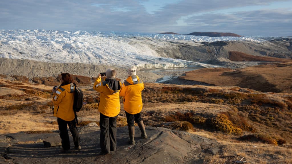 3 polar travelers stand in yellow jackets on a grassy knoll, overlooking a glacier during The Northwest Passage Canadian High Arctic voyage.