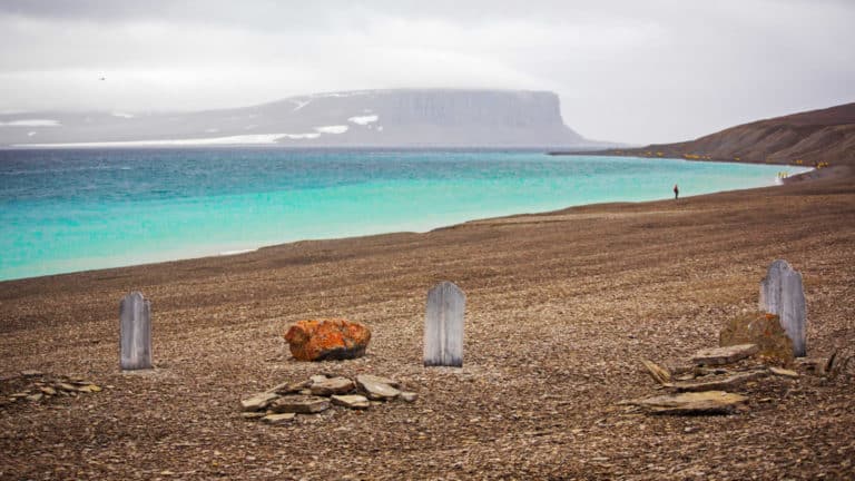 Beechey Island with 3 graves of explorers on a misty day with turquoise water along the brown, rocky shoreline, seen during The Northwest Passage Canadian High Arctic voyage.