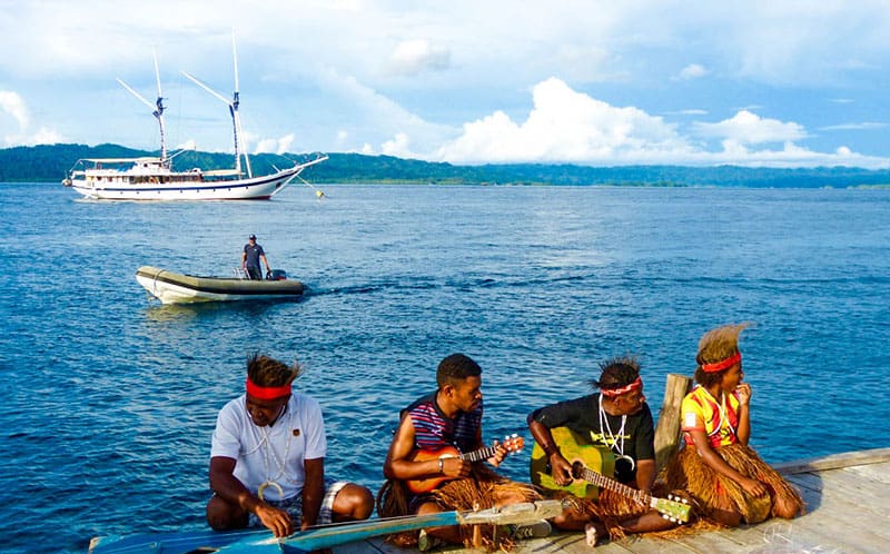 a group of Indonesian musicians sit on a dock in front of the ocean playing instruments, behind them floating in the water is an all white wooden small ship