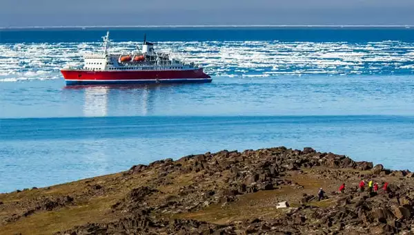 A red-hulled Arctic expedition ship in calm icy waters near a rocky brown shore.