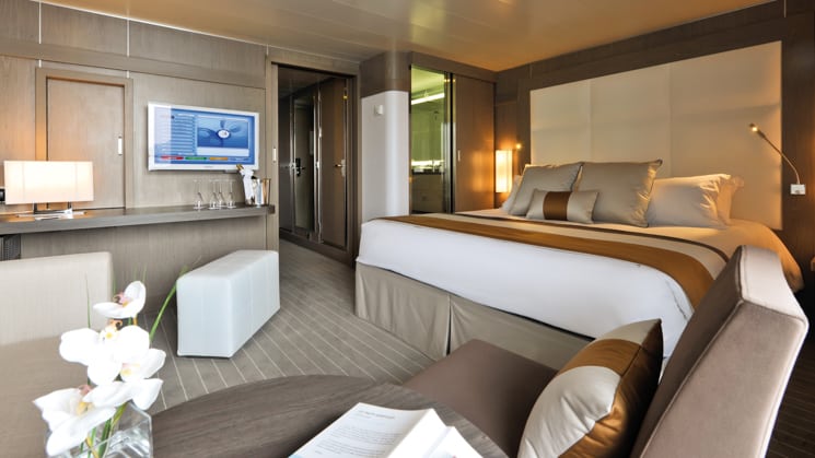 Deluxe Suite aboard L'Austral expedition ship, showing king bed, table, TV, chair & white-&-copper appointments.