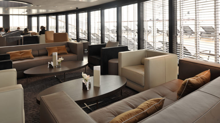Interior of L'Austral luxury French expedition ship, with tan couches, wooden coffee tables & wraparound windows.