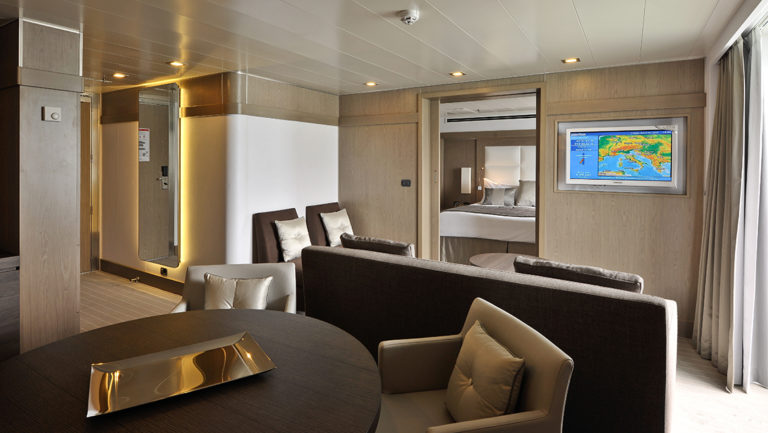 Owner's Suite aboard L'Austral french luxury ship, showing separate living room with couch, TV, table & chairs with view into bedroom.