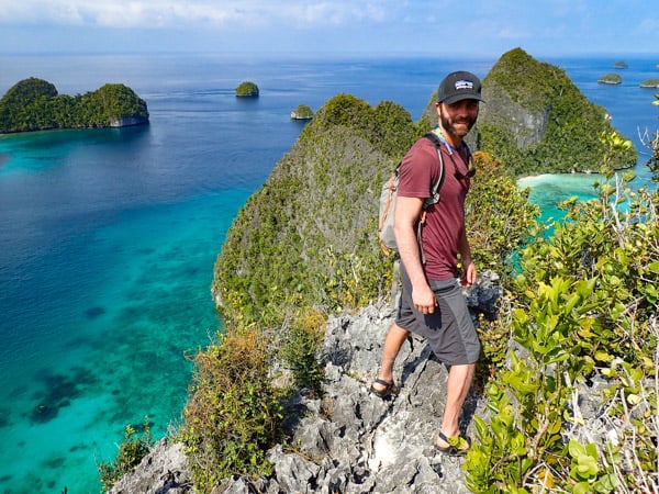 A male traveler stands at the very top of small island structures in Indonesia, blue sky day matches the teal waters, little green islands pop out of the water, seen as part of a small ship Indonesia cruise