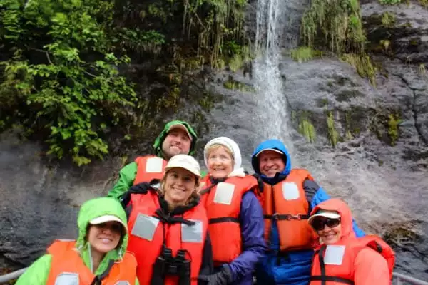 A family of 6 sit on a Kodiak infront of a waterfall, alll wearing orange life vests, as part of a daily activity abord an Alaska small ship cruise