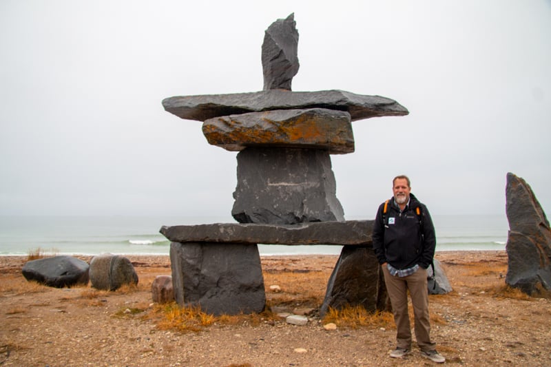 Todd Smith, AdventureSmith Explorations founder, stands in front of a large scale rock formation, also known as a cairn is a man-made pile of stones. The fall colors line the floor, yellow and orange, against the sea shore, on a foggy day.