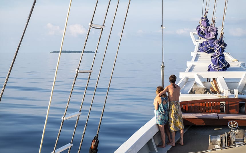 A couple embraces as they look towards the ocean horizon from the bow of a traditional Indonesian small ship. The sky matches the water in its blue hue almost to where you cannot tell where one ends and the other begins except for a single green island splitting the two. 