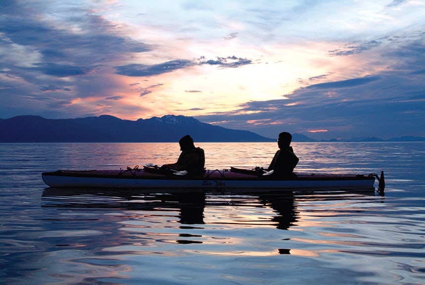 a couple sitting in a kayak is silhouetted against a pink, purple, yellow, and blue sunset on a alaska small ship cruise.