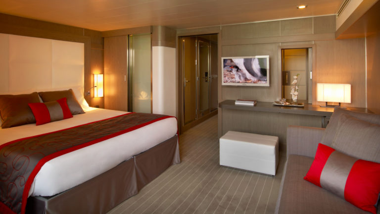 Deluxe Suite aboard Le Boreal expedition ship, showing king bed, table, TV, chair & white-&-copper appointments.