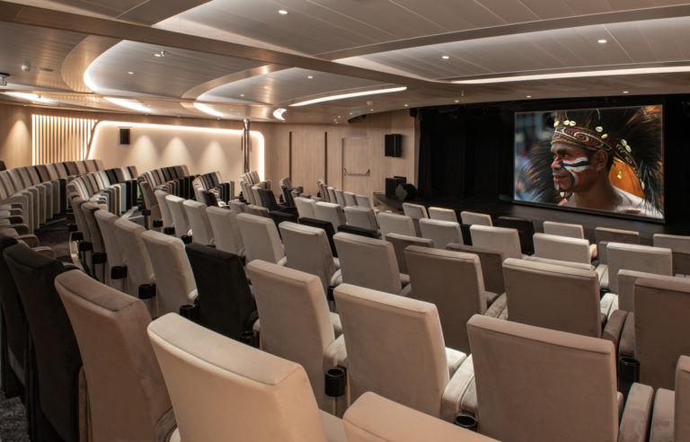 Theater aboard Le Champlain expedition ship, with rows of cushioned ivory seats, soft ceiling lighting & large TV screen showing indigenous culture.