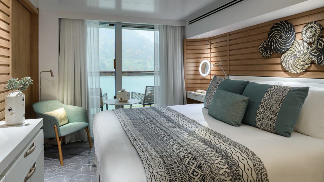 Prestige Suite aboard Le Champlain french small ship, with ethnic chic decor, white linen king bed, private balcony, wooden walls, desk & chair.